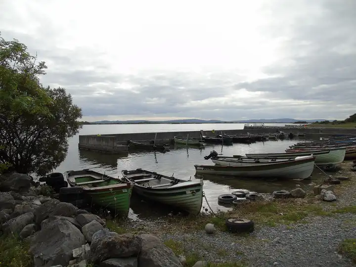 Lake boats moored in the small harbour at Annaghdown Pier, with the harbour wall, the open lower lake and distant hills under a grey sky.