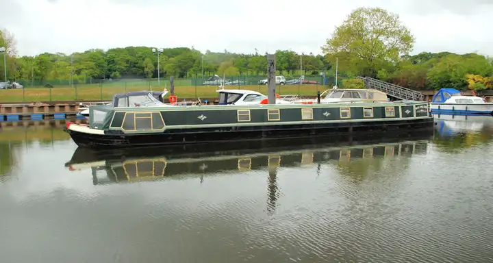 A long dark green narrowboat lies in the basin at Drumaheglis Marina, reflected in the calm water, with cruisers and the car park behind.