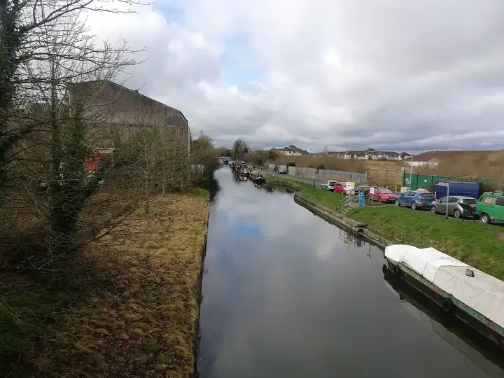 Het kanaal in Sallins gezien vanaf de brug, met een lange rij afgemeerde boten langs de oever, een afgedekte aak op de voorgrond en geparkeerde auto’s ernaast.