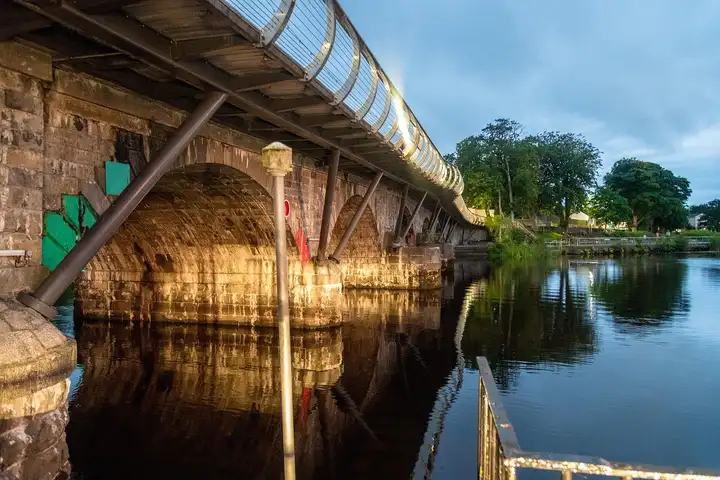 In de schemering branden lampen onder de bogen van de brug van Carrick-on-Shannon en langs de gebogen voetgangersbrug aan de zijkant, weerspiegeld in de kalme rivier.