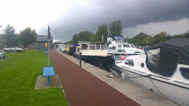Motorboten en een kanaalaak afgemeerd langs de kade van Shannon Harbour, naast een rood pad, bankjes en een grasveld onder een dreigende lucht.