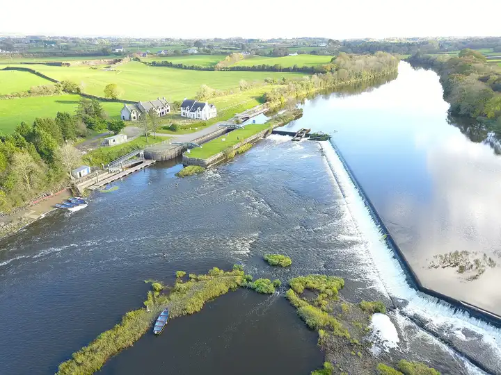 Aerial view of Carnroe on the Lower Bann, with the long white weir across the river, the lock cut and houses on the left and green fields beyond.