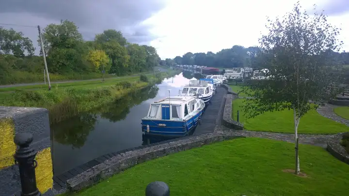 Cruisers moored in a line along a boardwalk in the canal harbour at Shannon Harbour, seen from a bridge, with a road along the far bank.