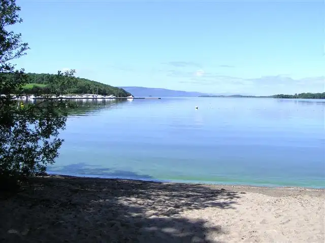 Clear shallow water laps a sandy beach in Muckross Bay, with a wooded headland and moored boats on the left and distant hills across the lough.