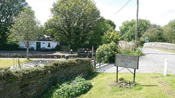A stone sign reading Coolnahay Bridge 26 stands among foxgloves beside the hump-backed bridge, with the lock gates and the whitewashed lock-keeper's cottage behind.