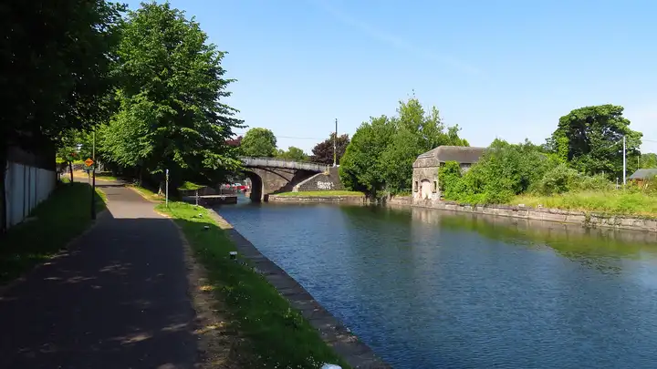 The wide canal east of Harbour Bridge in Mullingar reflects a cloudless sky, with a shaded path and trees on the left and an old stone building on the far bank.
