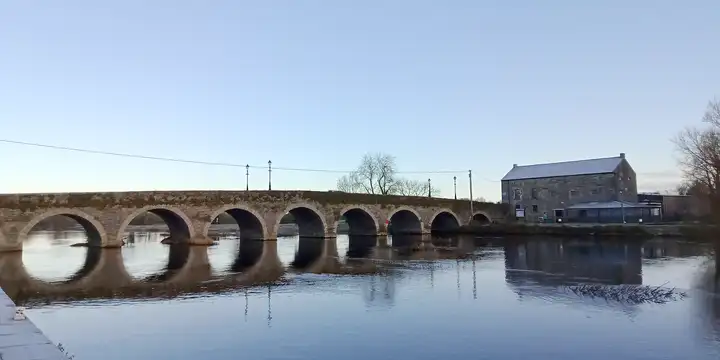 The granite arches of Gore’s Bridge mirrored in the calm River Barrow on a frosty morning, with an old stone store on the far bank.