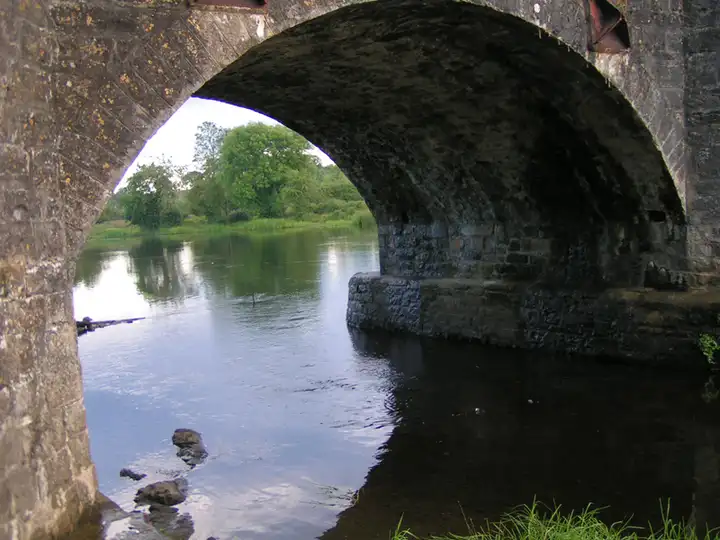 Seen from beneath an arch of Drumsna bridge, the Shannon flows calmly past a stone pier towards green fields and trees.