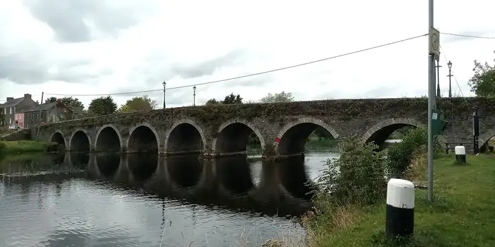 Gore’s Bridge gezien vanaf de oever op een bewolkte dag, met de bogen weerspiegeld in de Barrow en dorpshuizen aan het andere eind.