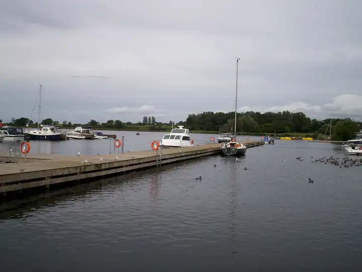 Ein Holzponton mit orangefarbenen Rettungsringen führt zu festgemachten Motorbooten und einer Segeljacht in der Kinnego Marina, unter grauem Himmel, auf dem Wasser Wasservögel.