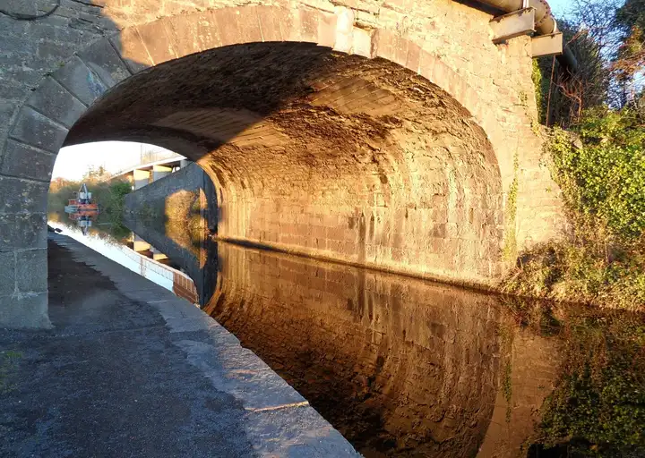 Low golden sunlight lights the stone arch of Louisa Bridge and its reflection in the still canal, with the towpath on the left and a small orange workboat beyond the arch.