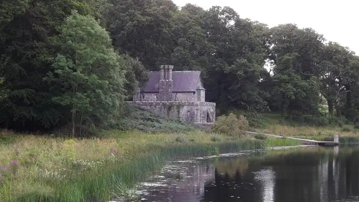 Het stenen boothuis van het landgoed Crom ligt onder hoge bomen aan het water, met rechts een lage steiger.