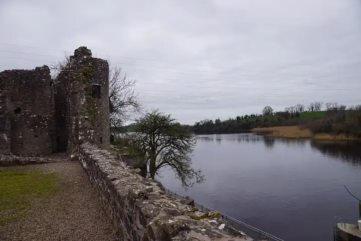 The ruined walls of Portora Castle stand above the wide, calm Erne at Portora, with reed beds and green fields on the far bank under a grey sky.