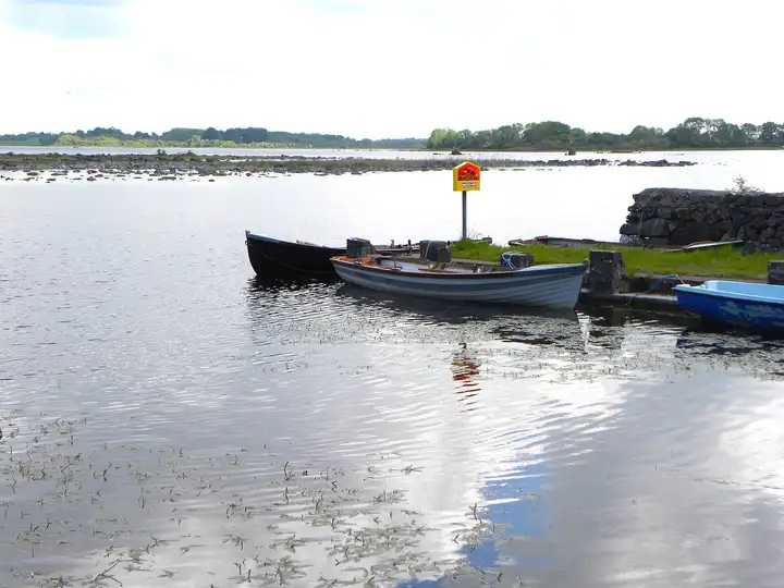 Two lake boats tied up at a low stone landing beside a yellow lifebuoy stand near Knockferry, with rocks and reeds in the shallow lake behind.