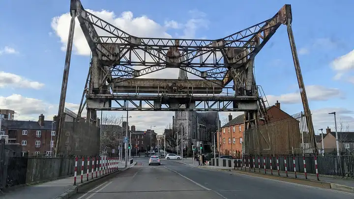 Das rostende Stahlgerüst der Klappbrücke an der Sheriff Street ragt über der Straße am Spencer Dock auf, dahinter Backsteinhäuser und ein Kirchturm.