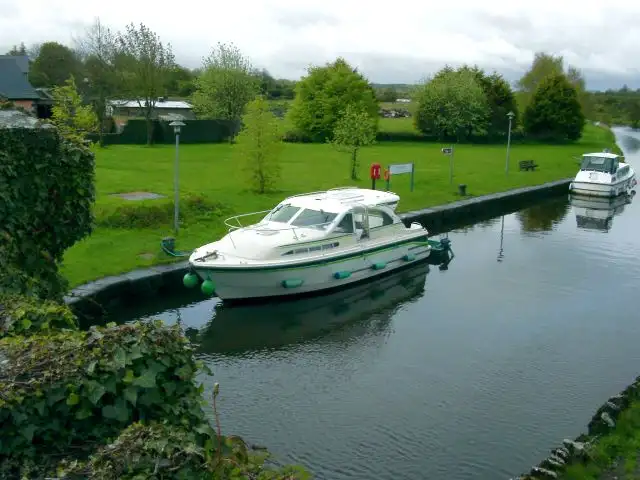 Von der Brücke in Leitrim aus gesehen liegt ein weißes Kajütboot am steinernen Kai neben einer Rasenfläche, weiter hinten ein zweites Boot.