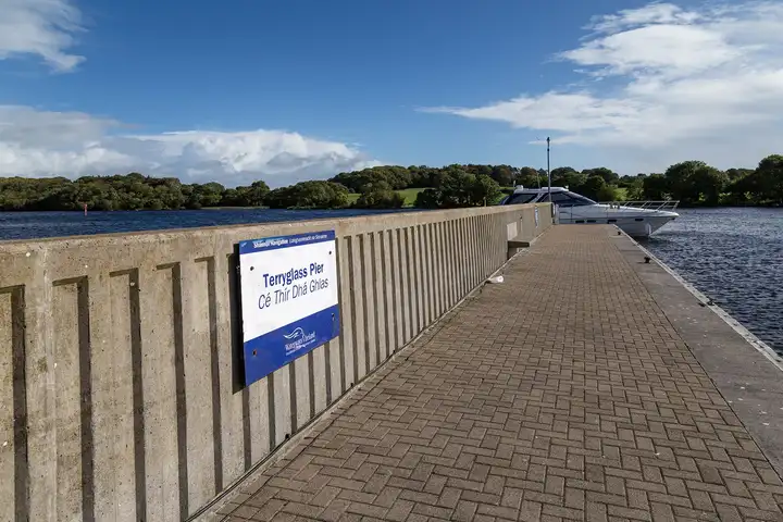 The paved breakwater of Terryglass Pier with its blue name sign, a cruiser moored at the end and a wooded shore across the water.
