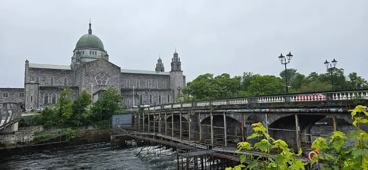 Salmon Weir Bridge in Galway on an overcast day, with rusty steel gantries over the rushing River Corrib below its arches and the green dome of Galway Cathedral behind.