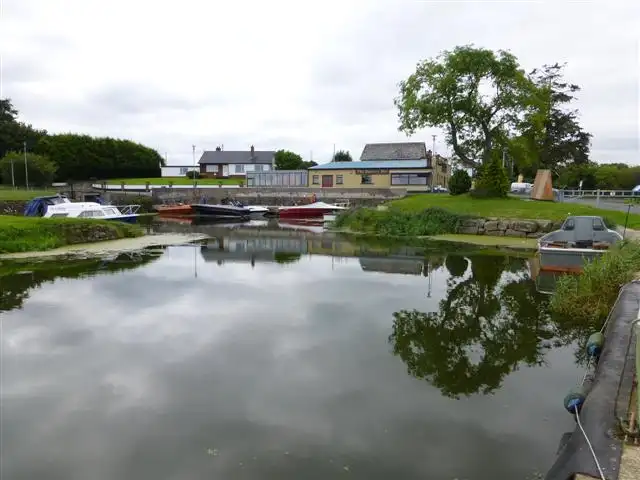 Le bassin intérieur du port de The Battery, avec de petits bateaux le long du quai, quelques maisons derrière et un grand arbre reflété dans l’eau immobile.