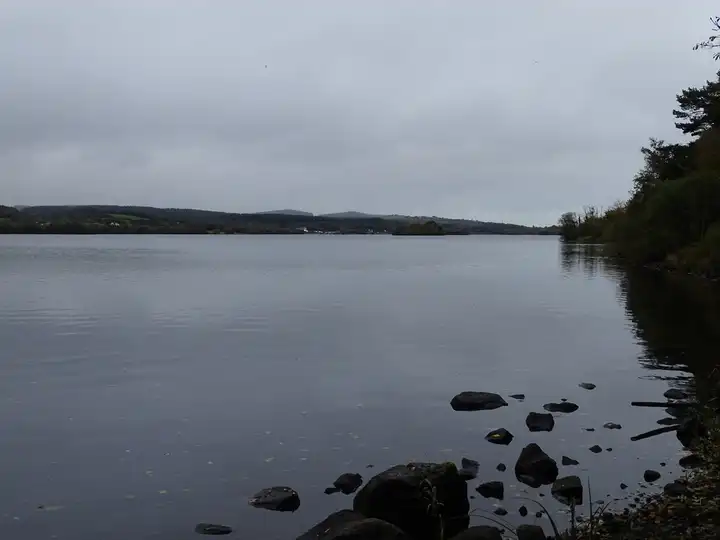 Dark rocks line the still, grey water below the wooded shore of Castle Caldwell, with hills fading into the overcast distance.
