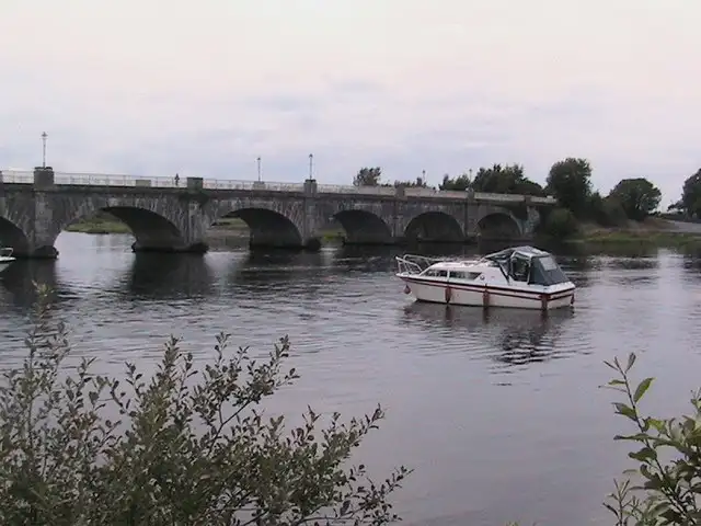 A small white cruiser on the Shannon just upstream of the stone arches of Banagher Bridge on a calm, grey day.