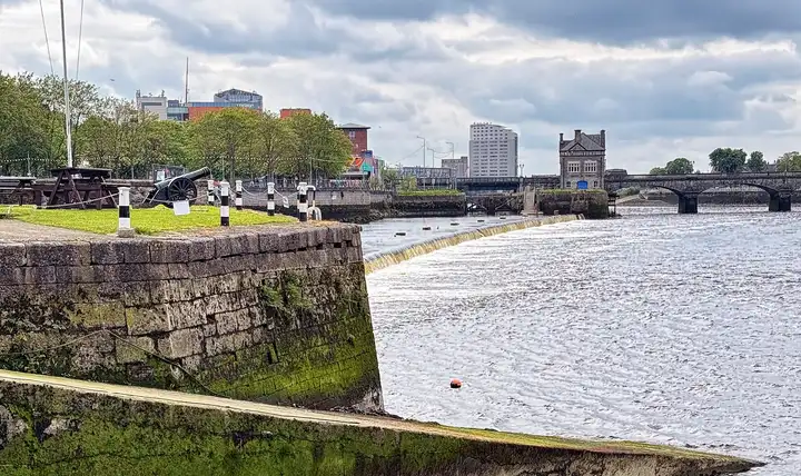 A low weir curves across the Shannon beside the lock walls, with an old cannon on the quay and Sarsfield Bridge in the distance.