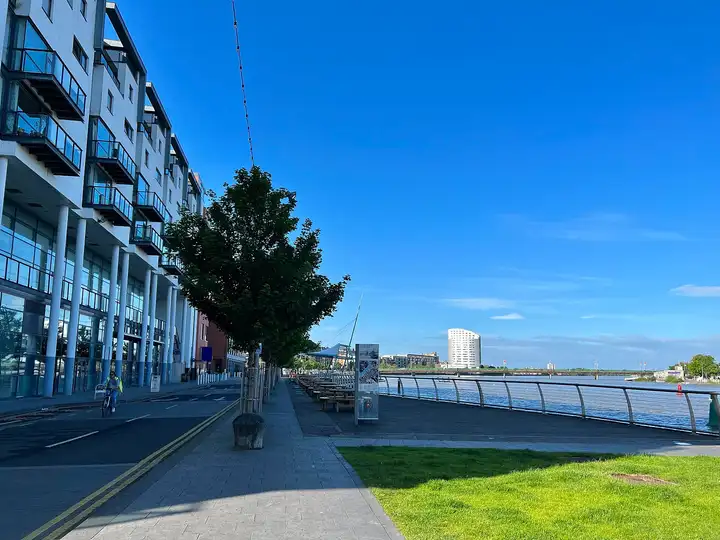 Eine sonnige Uferpromenade mit Picknicktischen verläuft in Limerick vor modernen Wohnblöcken am Shannon entlang.