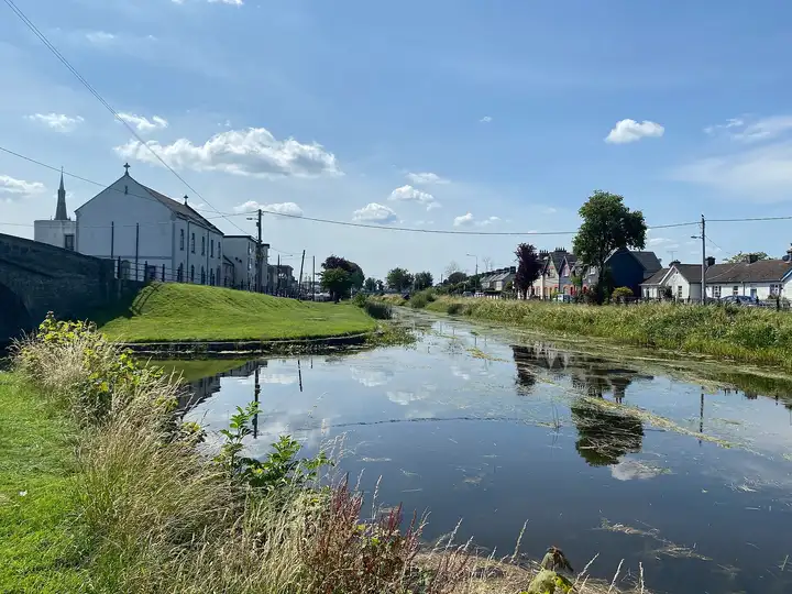 Der Grand Canal in Tullamore an einem Sommertag, mit einer weißen Kirche und Häusern am Wasser und Wolken, die sich auf der ruhigen Oberfläche spiegeln.