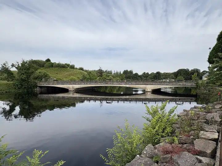The low concrete arches of Belleek Bridge span the River Erne and are mirrored in the calm water, with a green hill and trees behind and a rocky bank in front.