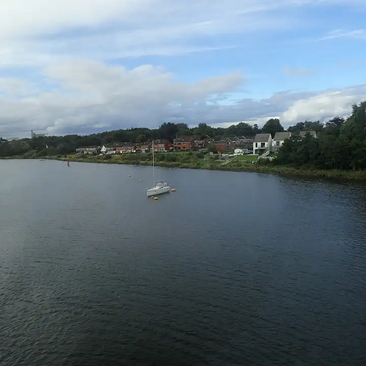 Un voilier blanc sur un corps-mort dans la large Bann, vu depuis le pont ferroviaire de Coleraine, avec une rangée de maisons sur l’autre rive.