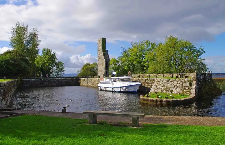A cruiser moored in Garrykennedy's old stone harbour beside the ruined castle tower, seen from the grassy bank.