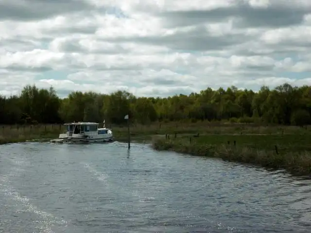 A small penichette cruiser on the Woodford River near Skelan Lock, passing a navigation marker between reed-fringed meadows and a line of trees.