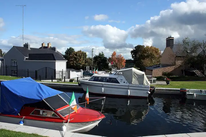 A red-and-white motorboat flying an Irish tricolour and a white cruiser lie moored in Abbeyshrule harbour under a blue sky, with houses and autumn trees along the quay.