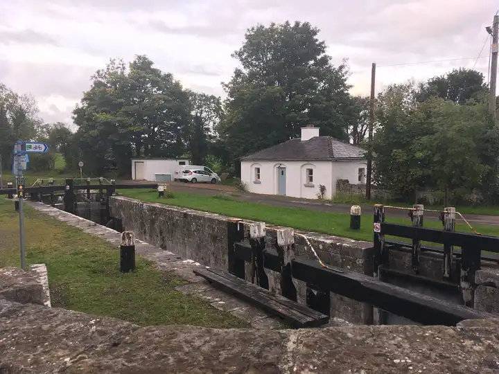 Seen over a stone parapet, the timber gates of Lock 43 at Killashee stand in front of the restored, whitewashed lock-keeper's cottage among tall trees.