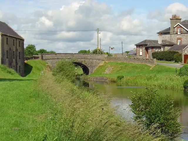 Der Kanal in Daingean führt unter einer Steinbrücke hindurch, links ein altes steinernes Lagerhaus, dahinter Häuser auf der Anhöhe.