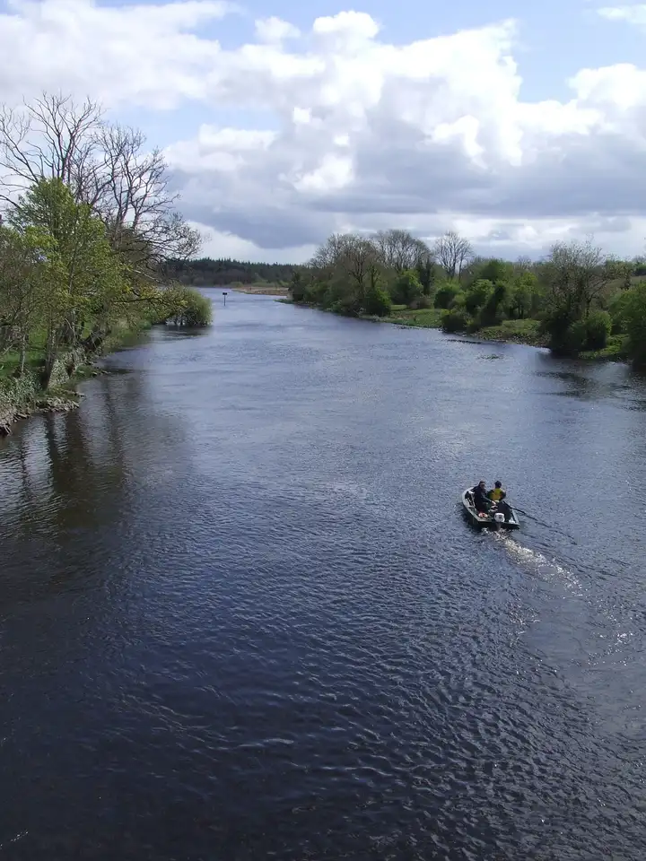 Looking downriver from Drumsna bridge, a small open boat with two anglers heads away along the broad Shannon between leafy banks.