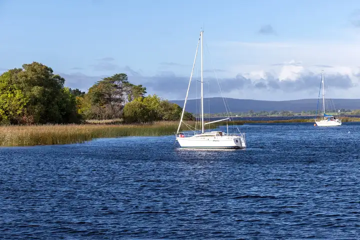 Two white sailing yachts at anchor on the blue water off Kilgarvan Quay, with reed beds, trees and distant hills behind.
