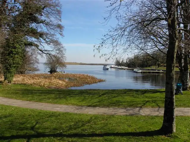 Hinter einer Rasenfläche und einem geschwungenen Weg auf Lusty Beg liegen kleine Motorboote an den Stegen einer ruhigen, schilfgesäumten Bucht unter klarem Himmel.