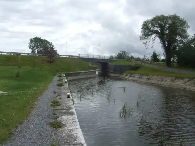 Bij Mosstown loopt het kanaal tussen een met steen beklede oever en een grazige helling naar de lage betonnen overspanning van Island Bridge onder een bewolkte lucht.