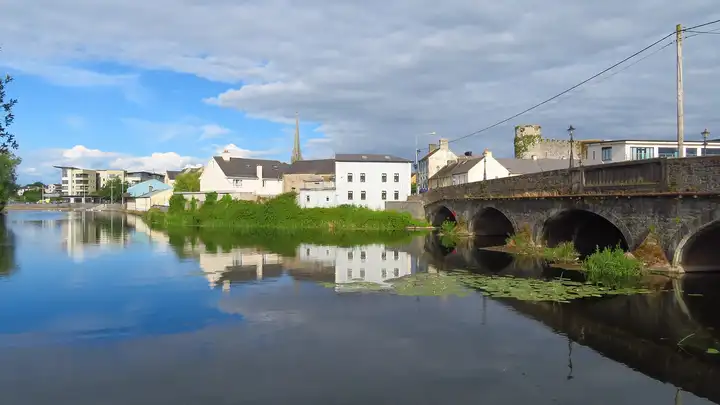 The low stone arches of Graiguecullen Bridge across the River Barrow at Carlow, with the ruined tower of Carlow Castle beyond and the town reflected in calm water.