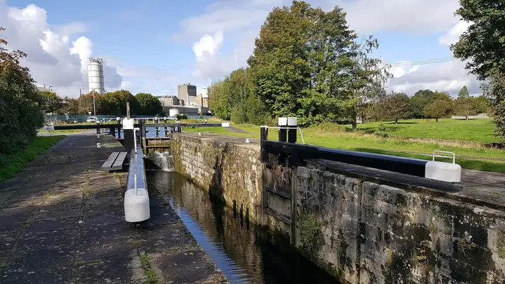 Die Steinkammer und die schwarz-weißen Schleusenbalken der Barrow Lock in Athy, hinter den Bäumen Industriegebäude und ein Silo.