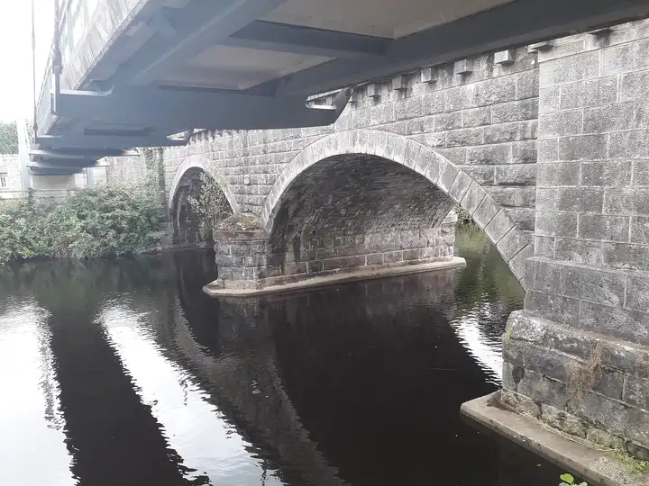 The dark water of the Woodford River flowing under the stone arches of Ballyconnell Bridge, seen from beneath the bridge deck.