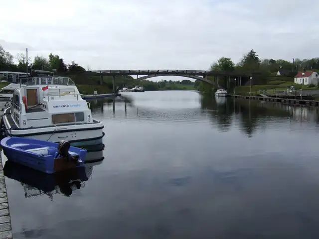A hire cruiser and a small blue boat moored at a pontoon in the foreground, with the arched Carry Bridge spanning the calm channel behind.