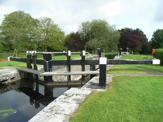 Les portes de bois noir et blanc et la passerelle de l’écluse 46 traversent le canal entre des pelouses soignées à Clondra, avec de grands arbres derrière.