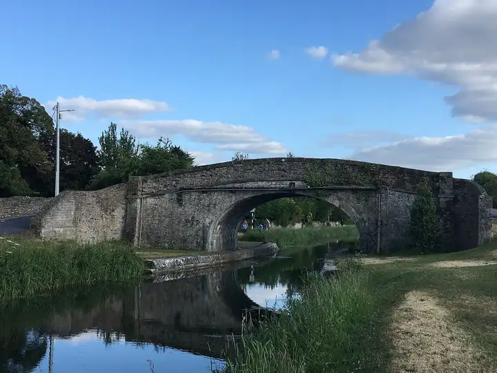 Die Abbey Bridge, eine Steinbogenbrücke über den Naas-Zweigkanal, spiegelt sich im ruhigen Wasser unter blauem Himmel mit weißen Wolken.