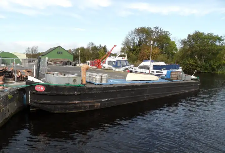 An old black canal barge marked 45M moored at a stone quay near Portumna Bridge, with cruisers and a red crane in the boatyard behind.