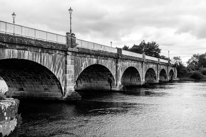 Black-and-white view of the broad stone arches of Banagher Bridge and the lamp posts on its parapet above the dark river.