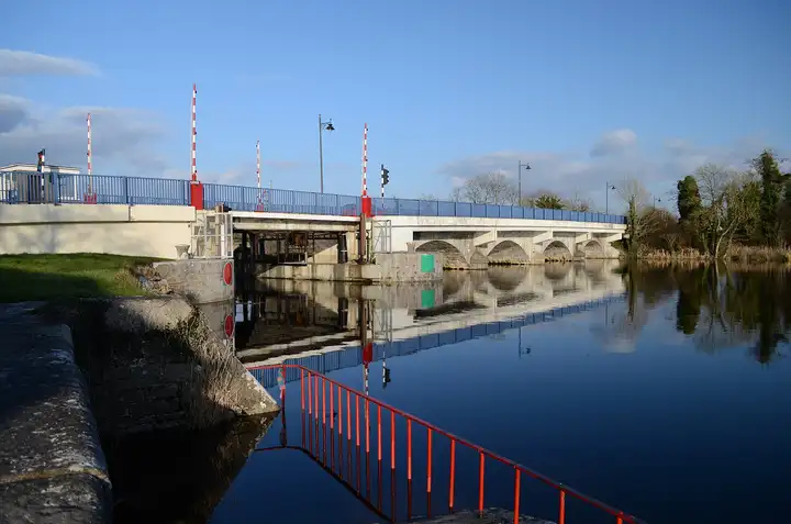 The N5 bridge at Tarmonbarry, with its lifting span and red-and-white barrier poles beside a row of stone arches, is reflected in still water under a blue sky.