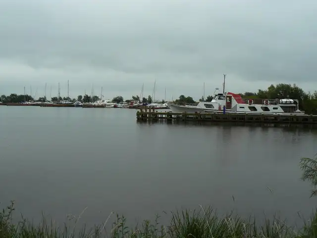 Een motorkruiser aan een houten steiger en een rij jachtmasten aan de overkant van het grijze, kalme water van de haven van Ballyronan op een bewolkte dag.