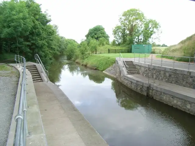 Seen from beneath the bypass bridge west of Enfield, the canal runs between new stone banks with steps and handrails towards green fields and a small green hut.